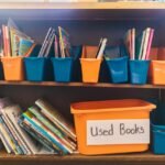Colorful bins filled with children's books on a wooden shelf, labeled as 'Used Books'.